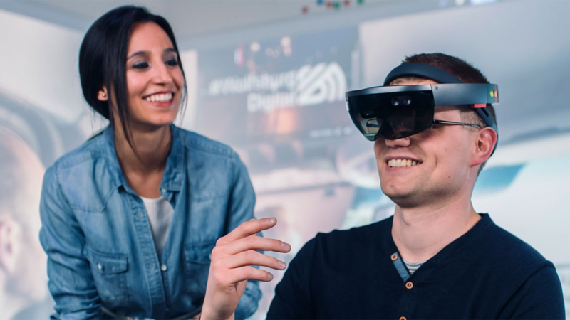 Junge Frau steht in einem IT-Lab neben einem jungen Mann, der eine VR Brille trägt