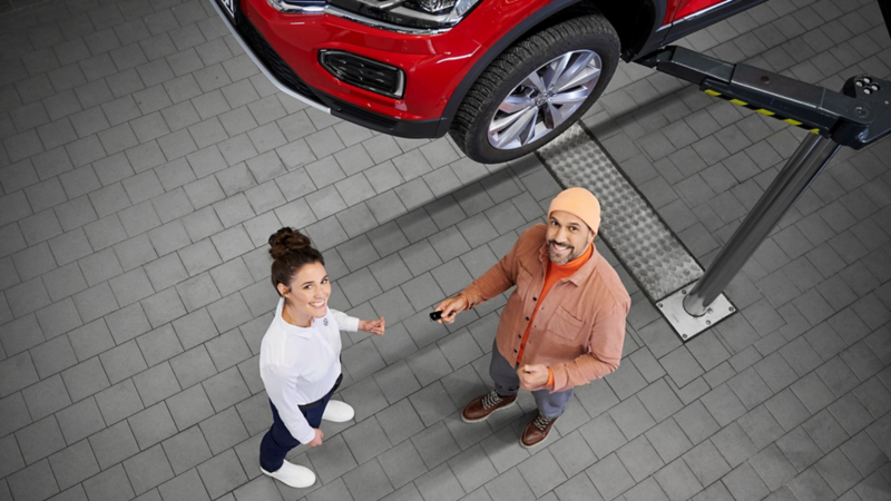 Na podnośniku widoczny jest czerwony Volkswagen. Pod nim stoi mężczyzna oraz pracownica serwisu VW.