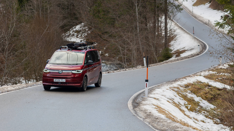 Red Volkswagen Multivan with a roof rack driving along a winding, snow-lined road through a forest. The scene conveys a sense of adventure and calm.