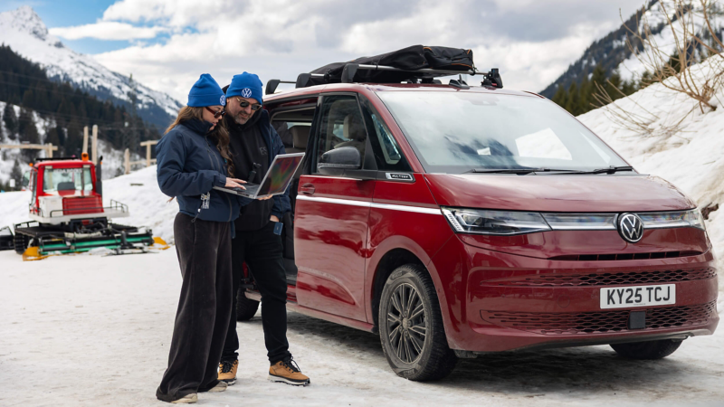 A couple in blue beanies stand beside a red Volkswagen Multivan on a snowy mountain road, sharing a laptop. The scene conveys adventure and exploration.