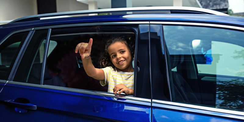 Bereit für einen Ausflug – Sophie schaut aus dem VW Tiguan heraus und winkt mit ihrem Kuscheltier Ted Turbo