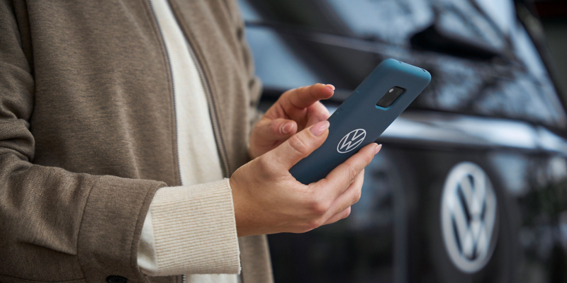 Nahaufnahme von Händen mit Smartphone in VW Hülle, im Hintergrund ein VW Auto