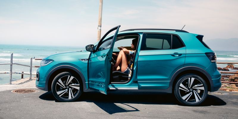 Een turquoise VW T-Cross met geopende deur aan de kant van de bestuurder op een parkeerplaats met uitzicht op zee
