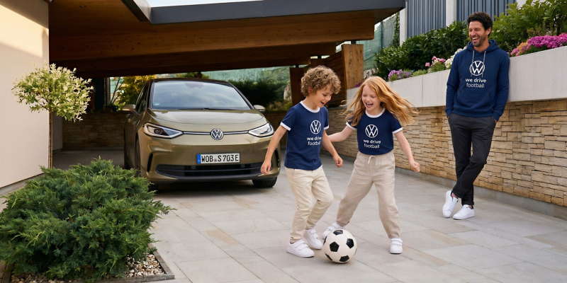 Kinder mit blauen T-Shirts aus dem Fußball Drop spielen Fußball in der Einfahrt, Mann im Hoodie schaut zu, ID.3 unter Carport
