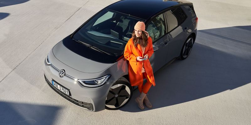 Eine Frau mit Smartphone in der Hand lehnt sich an ihren grauen VW ID.3 an