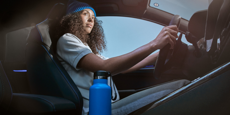 Frau mit blauer Mütze aus der R-Kollektion sitzt auf Fahrersitz eines VW Autos, in der Mittelkonsole blaue Trinkflasche