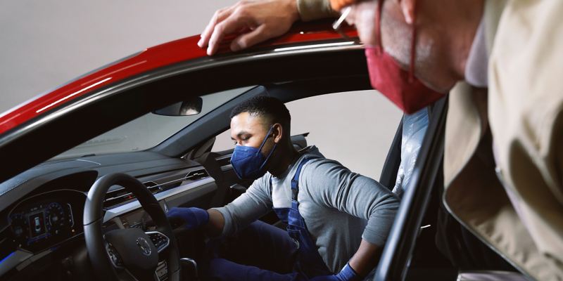 Ein VW Servicemitarbeiter mit Atemschutzmaske prüft den Innenraum eines VW während Corona, der Kunde steht neben dem Auto