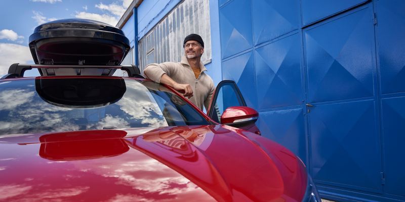 Ein Fahrer steht an seinem roten Taigo mit VW Dachbox – Volkswagen Transport