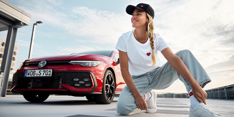 Woman with VW GTI cap and GTI T-shirt from VW lifestyle collection next to Volkswagen car