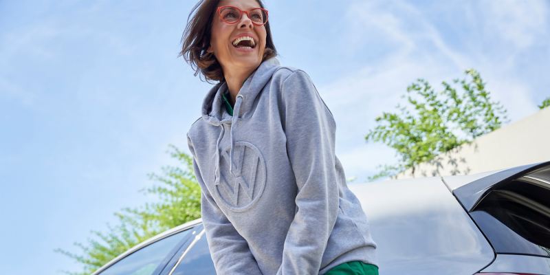 Eine lachende Frau, bekleidet mit einem VW Sweatshirt, vor einem VW Auto – Volkswagen Zubehör