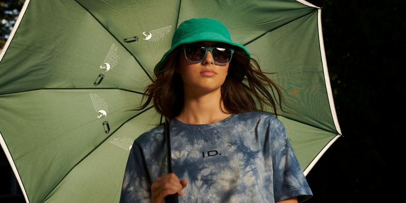 Eine Frau mit Sonnenbrille und Batik-T-Shirt bekleidet, hält einen Regenschirm aus der ID. Buzz Kollektion