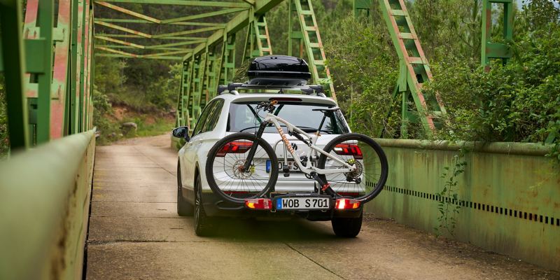 VW Tiguan met een dakkoffer en een fietsendrager van VW-accessoires incl. fiets rijdt over een brug