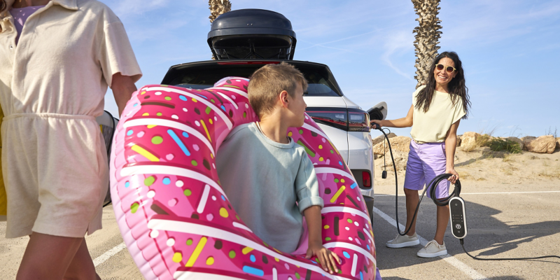 Zwei Frauen und ein Kind mit Strandspielzeug neben VW Auto mit Wallbox