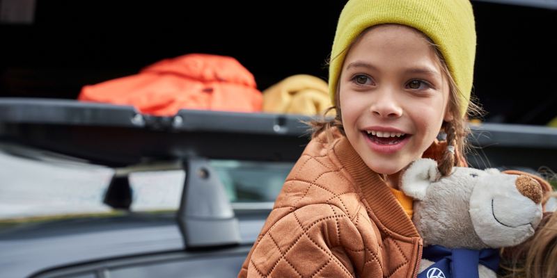 Ein Mädchen mit Kuschelbär auf dem Arm, im Hintergrund eine geöffnete VW Zubehör Dachbox