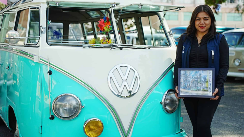 Mujer muestra cuadro conmemorativo junto a Combi Volkswagen clásica en evento Treffen.