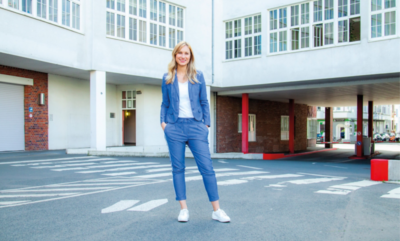 Eine lächelnde Frau im blauen Anzug steht im Hof eines VW Standorts