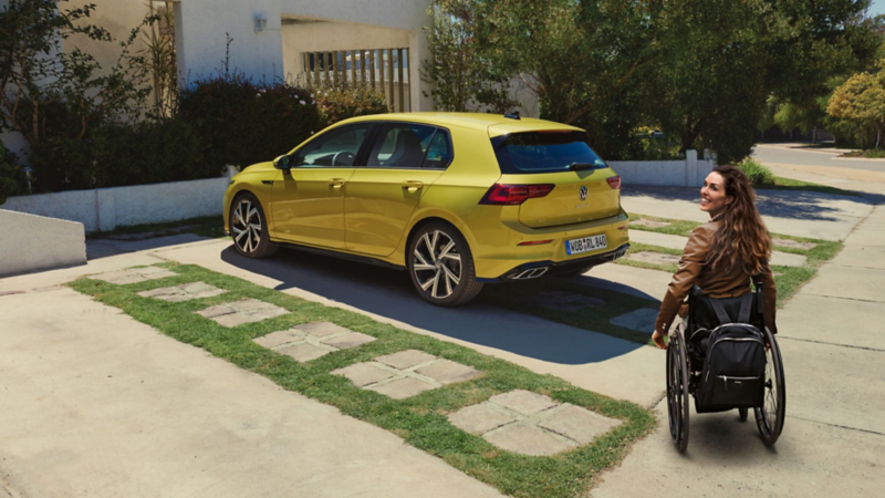 Devant une maison, une Volkswagen Golf de couleur jaune vif est stationnée. Une femme souriante est assise dans un fauteuil roulant à proximité