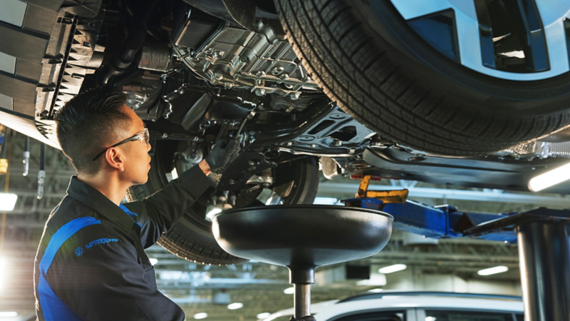 A VW service technician stands underneath a Volkswagen that is on a car lift, draining the old oil out and into a catch pan.