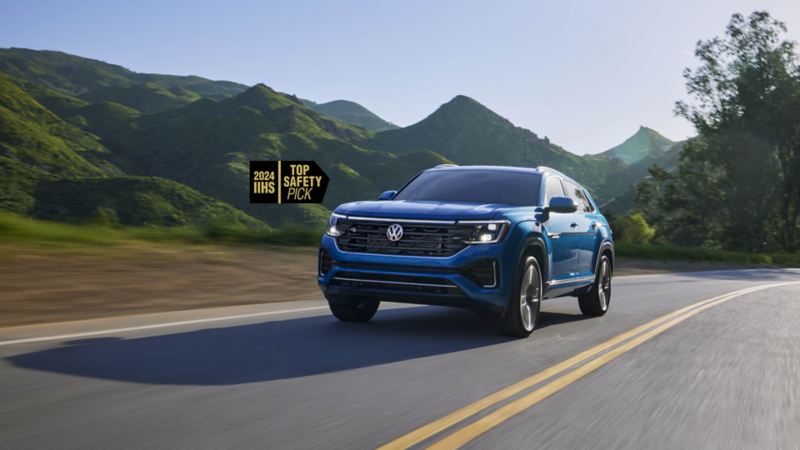 A blue 2024 Cross Sport, an IIHS "Top Safety Pick," is driving on a road with green hills in the background.
