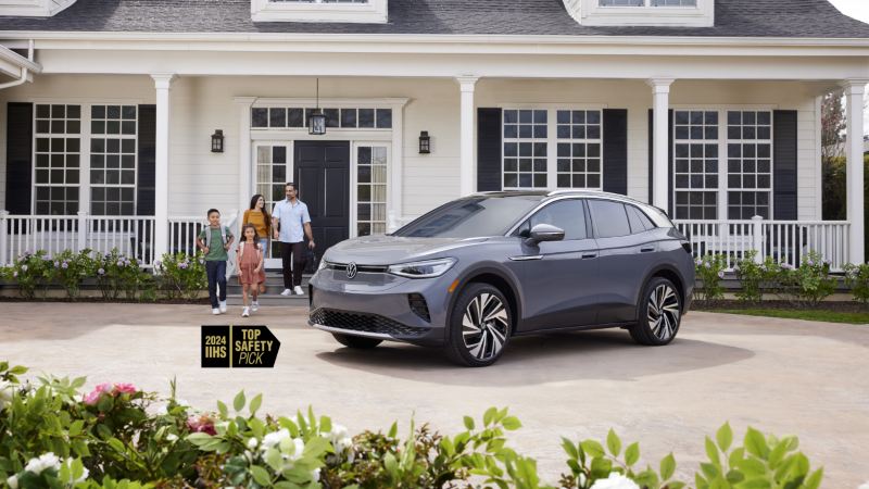 The award-winning 2024 ID.4, an IIHS "Top Safety Pick," is parked in front of a white house, and two adults and two children are walking on the driveway towards the vehicle.