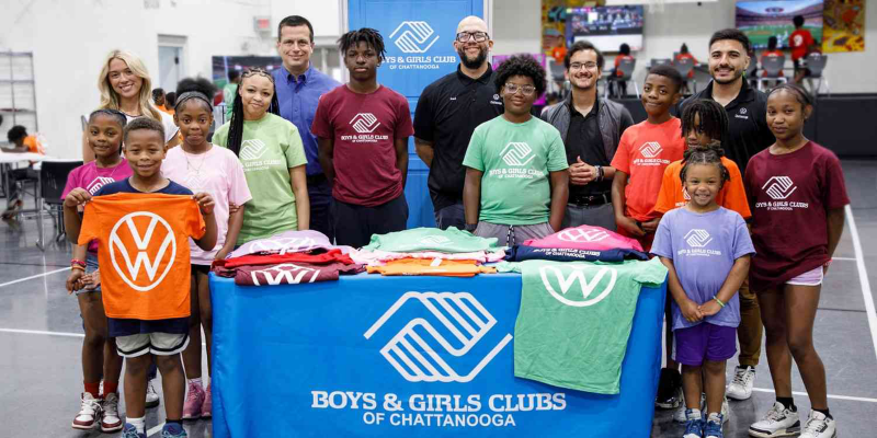 A group of children and adults pose behind a table with a "Boys & Girls Clubs of Chattanooga" banner, featuring colorful t-shirts and a gymnasium background.