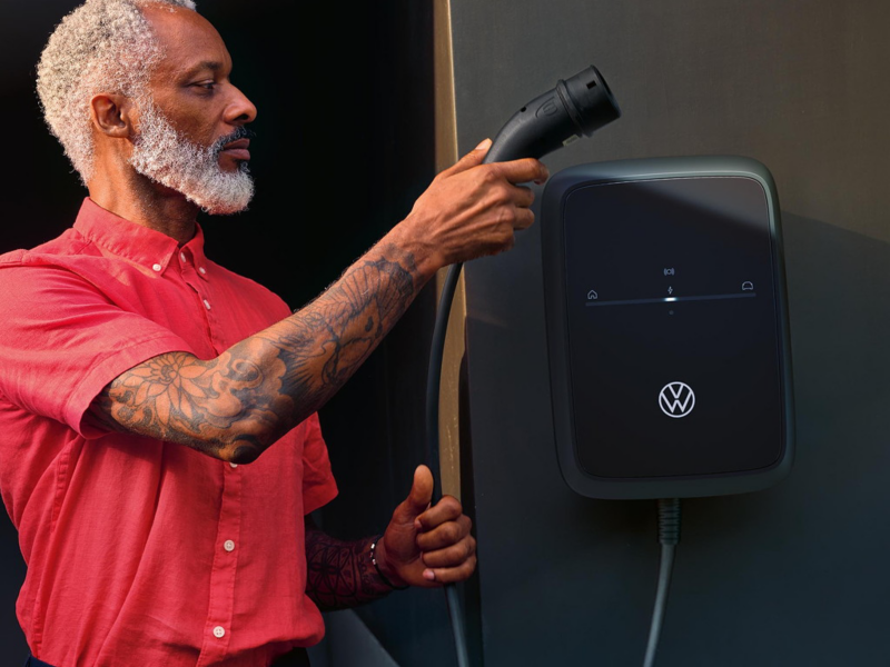 Mann mit rotem Shirt hält einen Ladestecker und steht vor einer an der Wand montierten VW Wallbox