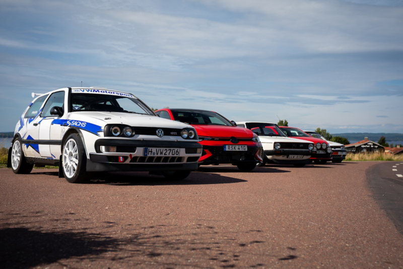 Fem VW Golf GTI från olika generationer uppställda på rad utomhus, från en vit rallybyggd Golf i förgrunden till äldre klassiska modeller och en modern röd Golf, med sjö och landsbygd i bakgrunden.