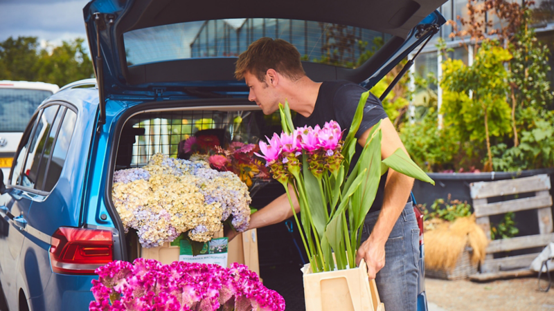 Un homme charge le coffre d'un Touran bleu avec des fleurs devant une jardinerie.