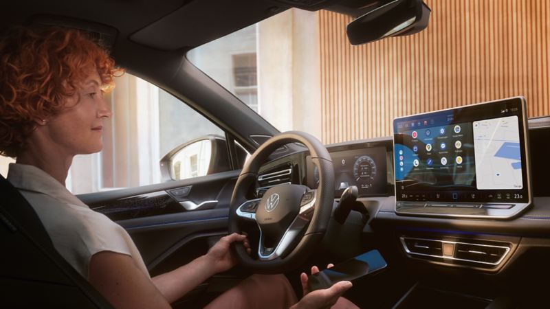 A woman is sitting at the steering wheel of the VW Tiguan interacting with the optional App-Connect and her smartphone.