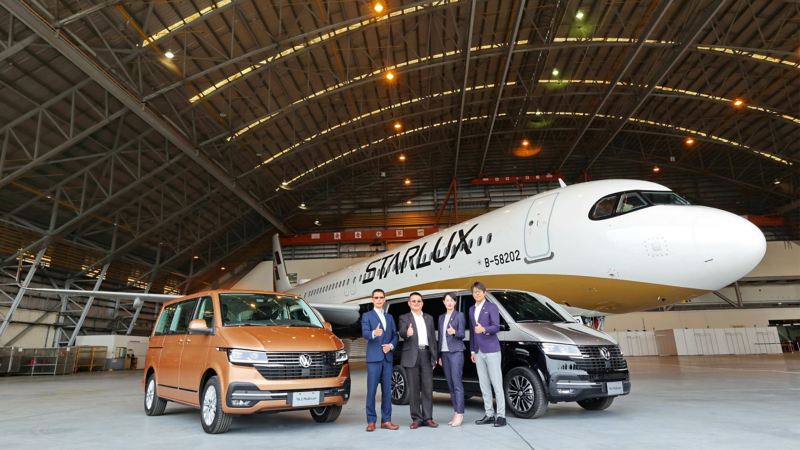 倉庫裡有兩台T6.1 Multivan，一台古銅金色、另一台棕黑雙色，兩車間站著四個人比讚，正後方有一架飛機