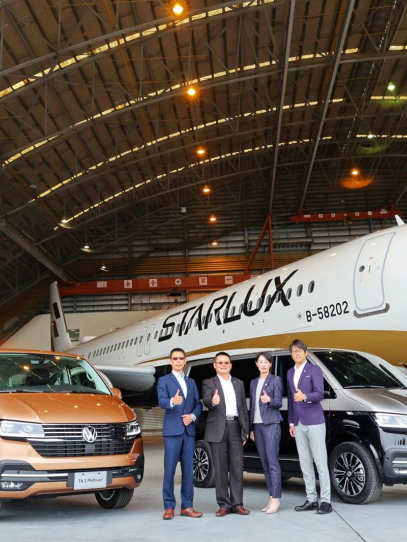 倉庫裡有兩台T6.1 Multivan，一台古銅金色、另一台棕黑雙色，兩車間站著四個人比讚，正後方有一架飛機