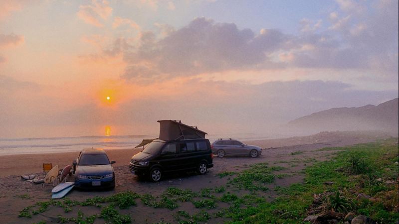 黑色California與另外兩台wagon車款停在沙灘上，車旁放著衝浪板，遠方可見夕陽掛在天空