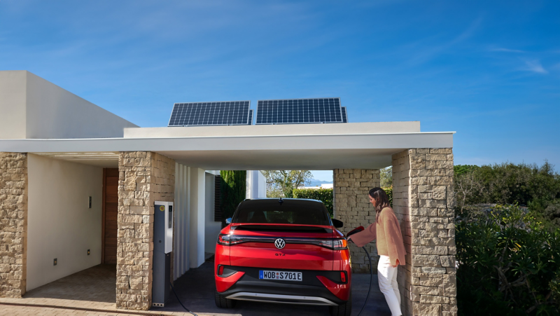 Ein VW ID.5 GTX lädt unter einem Carport mit Solarpanels, eine Frau steht daneben