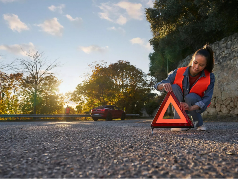 Mujer con chaleco naranja colocando un triángulo de peligro en la carretera.