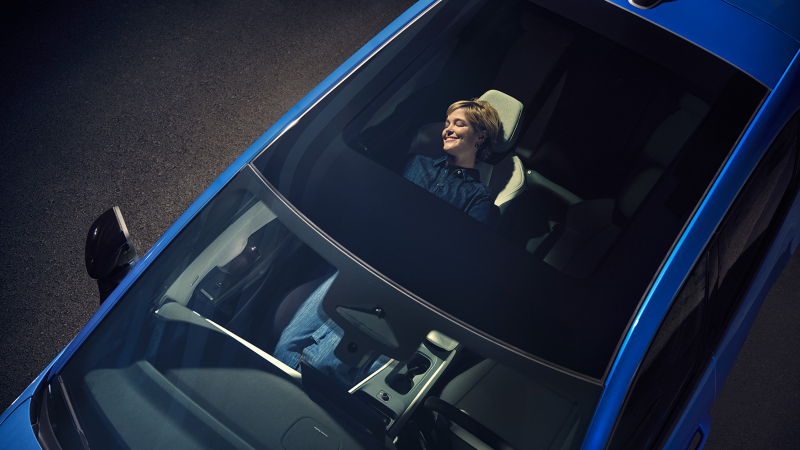 Blick von oben durch das Panoramadach eines blauen VW ID. Polo Style. Auf dem zurückgelehnten Beifahrersitz entspannt eine Frau.