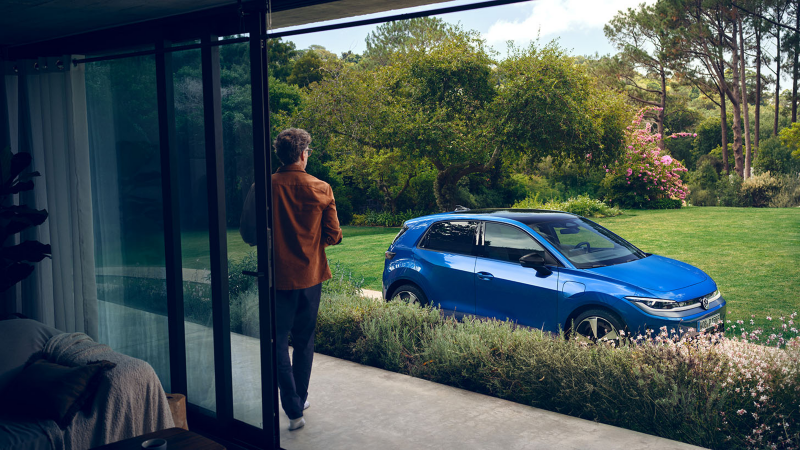 Blick durch eine offene Terrassentür in einen grünen Garten, in der Auffahrt steht ein blauer VW ID. Polo Style.