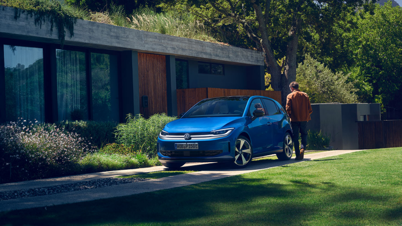 Ein blauer VW ID. Polo Style parkt vor einem begrünten Wohnhaus, ein Mann geht darauf zu.