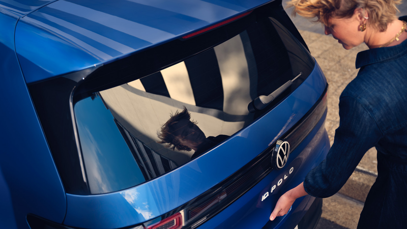 Eine Frau öffnet die Kofferraumklappe eines blauen VW ID. Polo Style.