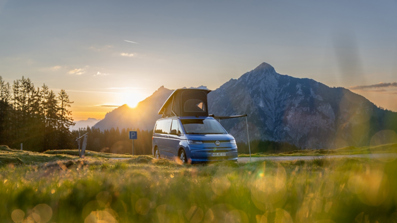 Un Volkswagen California bleue, toit relevé, se trouve devant un panorama montagneux au crépuscule ; la scène est baignée d'une lumière dorée.