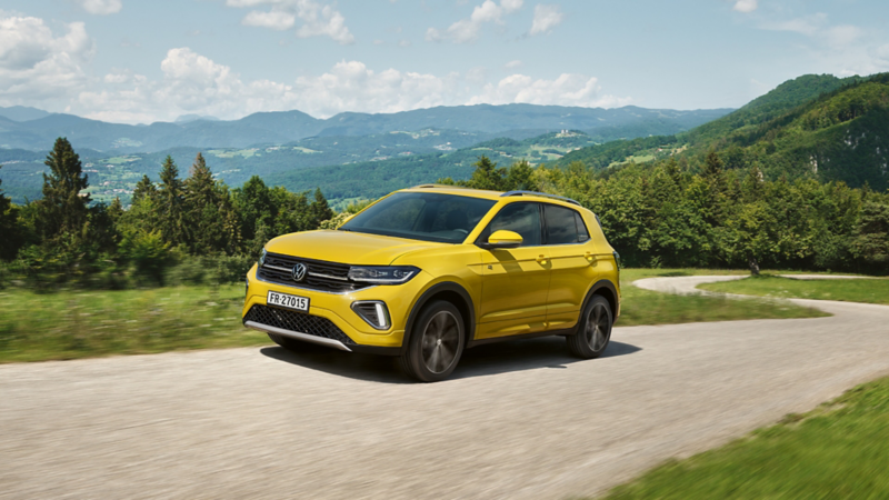 Volkswagen T-Cross in Gelb fährt dynamisch auf einer kurvigen Landstrasse mit grüner Hügellandschaft im Hintergrund.