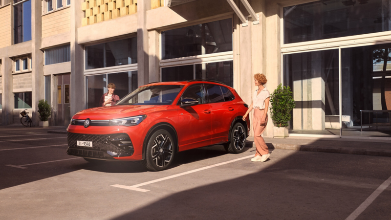 Volkswagen Tiguan rosso parcheggiato in un parcheggio urbano, con una donna accanto e un giovane sullo sfondo.