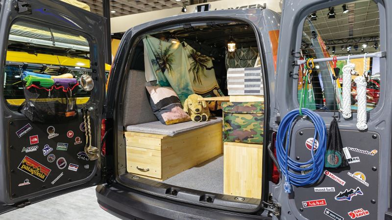 灰色Caddy Van改裝成VanLife露營車，尾門向兩側開啟可看到門上掛滿戶外運動小道具，後廂加上木箱堆疊成的座位、收納空間及工作平台