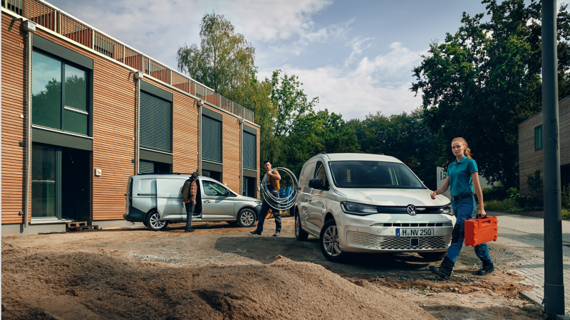 女性維修人員手提工具箱經過香草白色的Caddy Cargo車頭，男性維修人員搬著工具站在車尾