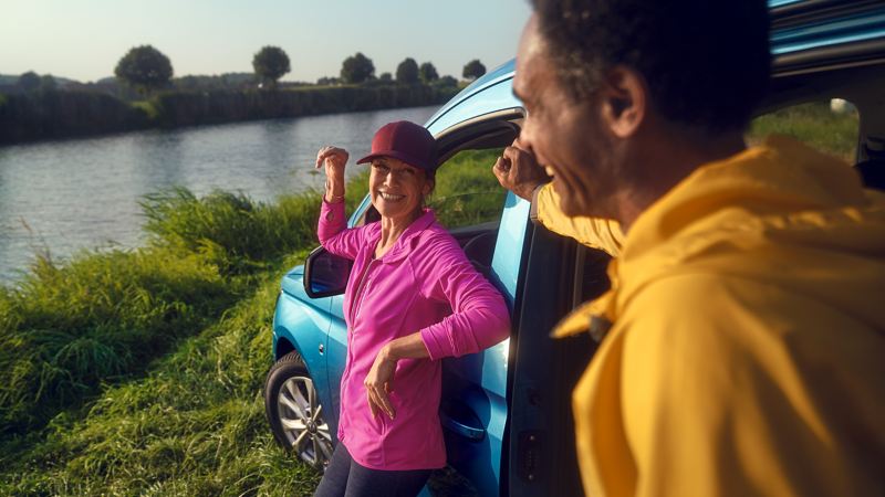 Caddy Maxi情境照-海水藍色車款前方站一位穿桃紅色的女子以及黃色衣服的男子，在河旁邊開心聊天