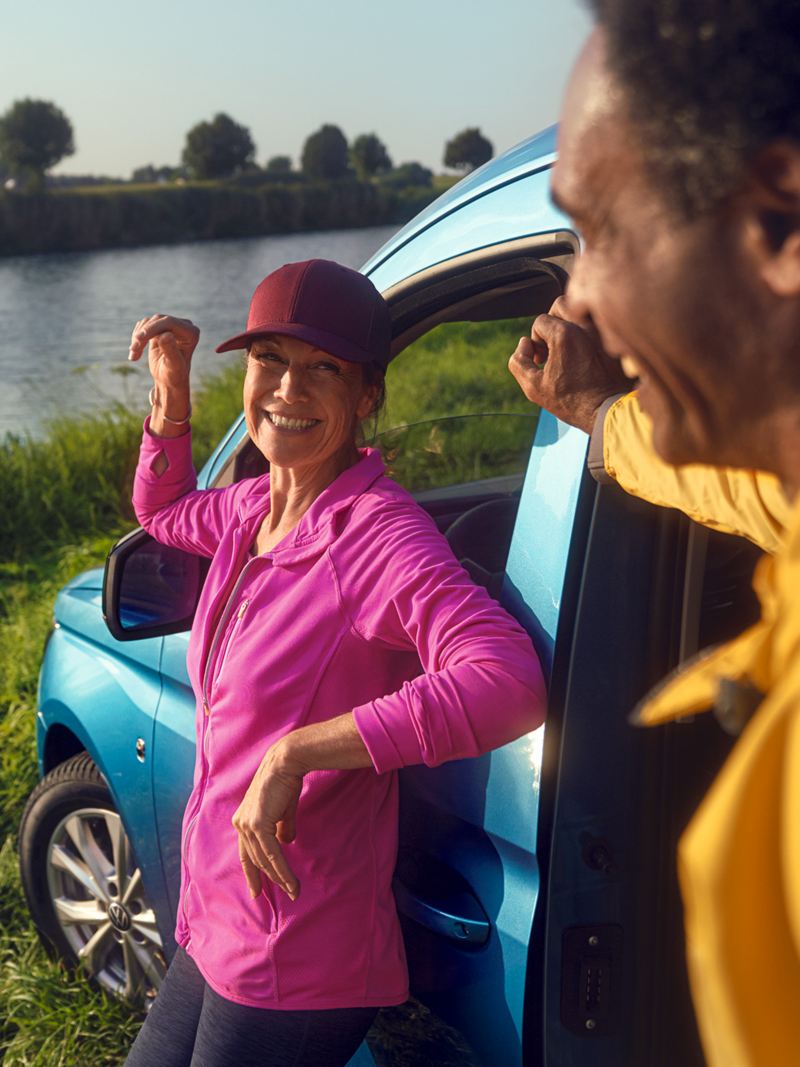 Caddy Maxi情境照-海水藍色車款前方站一位穿桃紅色的女子以及黃色衣服的男子，在河旁邊開心聊天