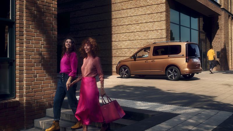 Caddy Maxi情境照-古銅金色車款停在街上，前面有兩個女子穿桃紅色上衣配牛仔褲以及蓮藕色上衣配淘紅色長裙
