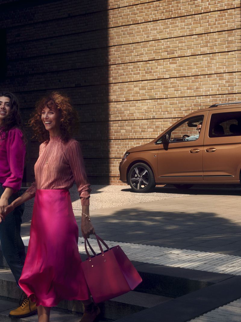 Caddy Maxi情境照-古銅金色車款停在街上，前面有兩個女子穿桃紅色上衣配牛仔褲以及蓮藕色上衣配淘紅色長裙