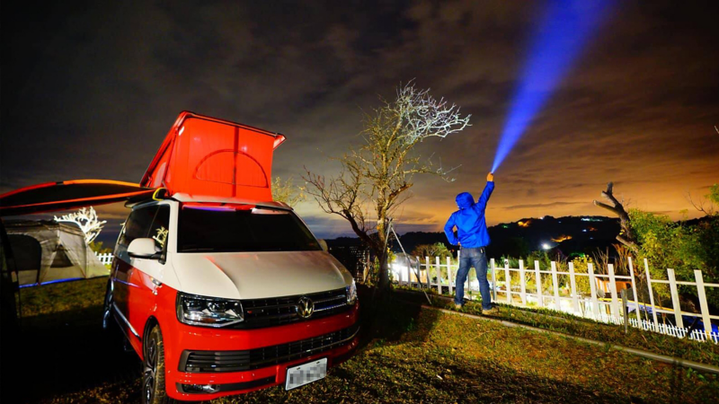 車主背對鏡頭站在紅白雙色California旁，一手插腰、另一手拿著強力手電筒開燈指向天空，看起來就像一道光束從手上往天空射出