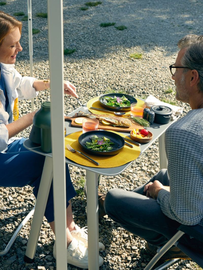 T6.1 California使用情境照，一對夫婦使用隨車附贈的桌椅在車側帳拉開下的戶外空間用餐