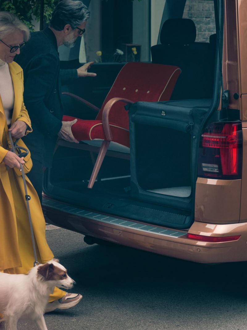 一位身穿黃色大衣的女性牽著小狗，站在古銅金的Multivan的車尾準備讓小狗上車